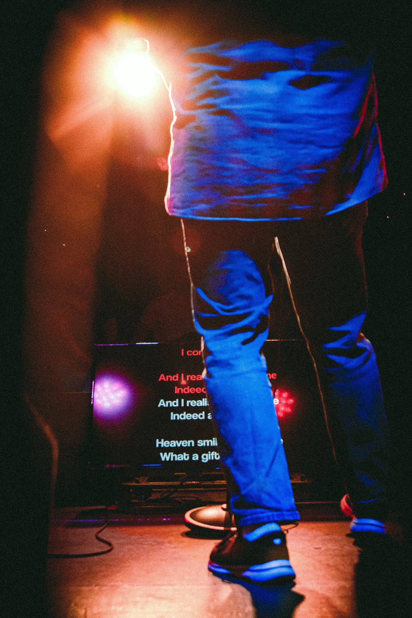 Person standing on stage in blue jeans and sneakers illuminated by bright light with a screen showing partially visible song lyrics in the background. bandkaroke.live