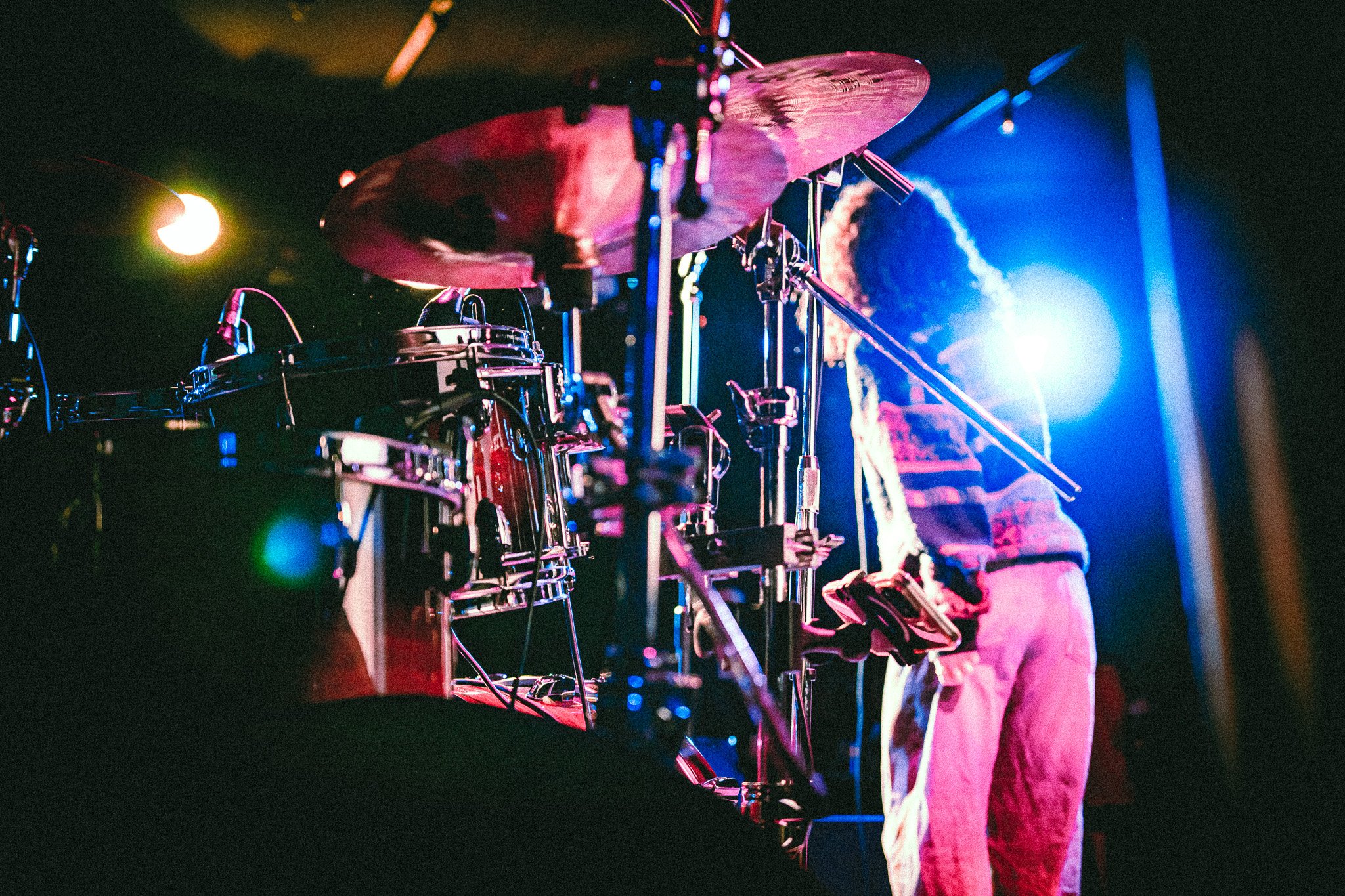Side view of a drum set on stage with a musician playing guitar under colorful stage lighting. bandkaroke.live