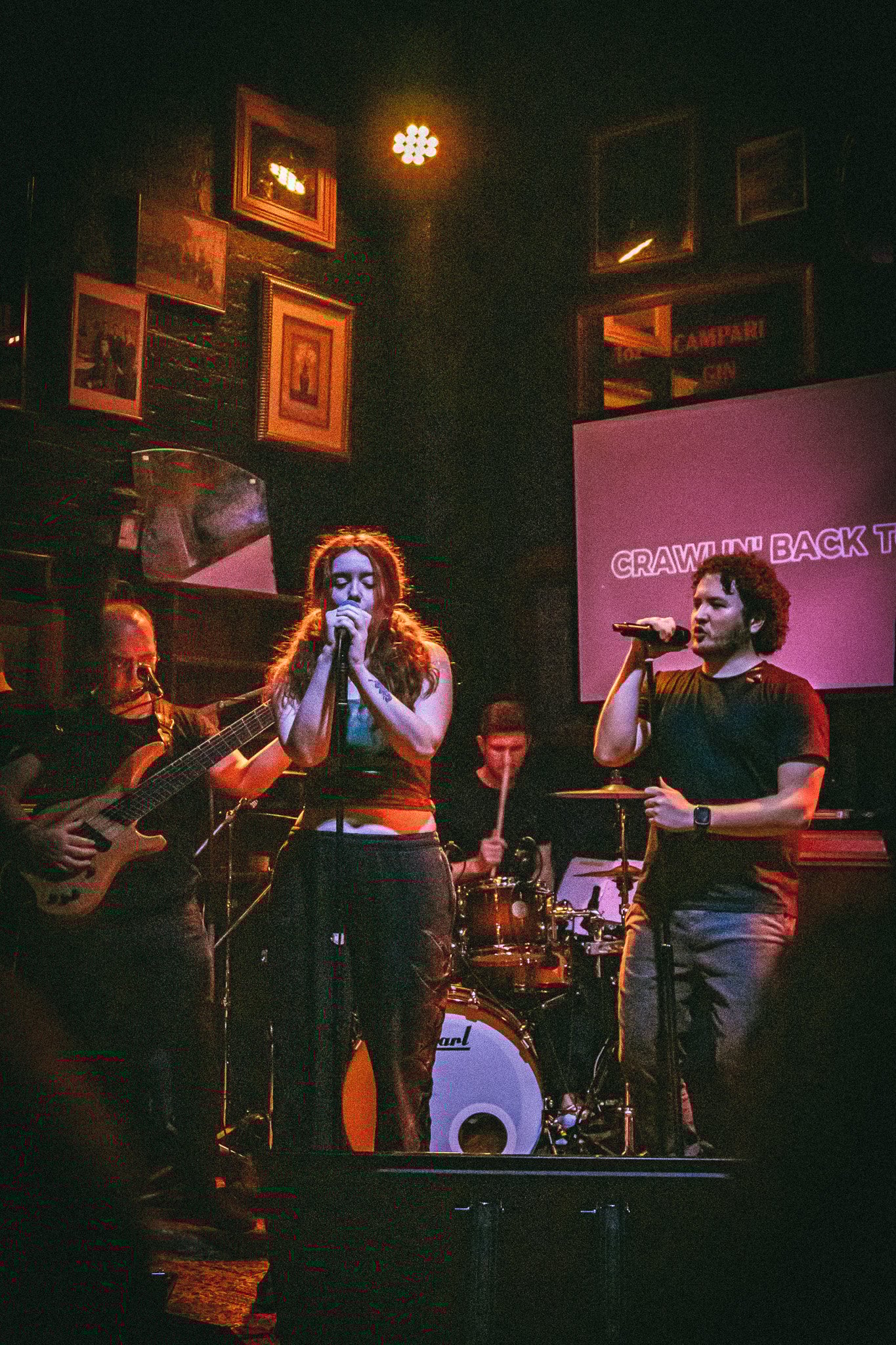 Four-member band performing on stage with two vocalists, a guitarist, and a drummer under warm lighting. bandkaroke.live