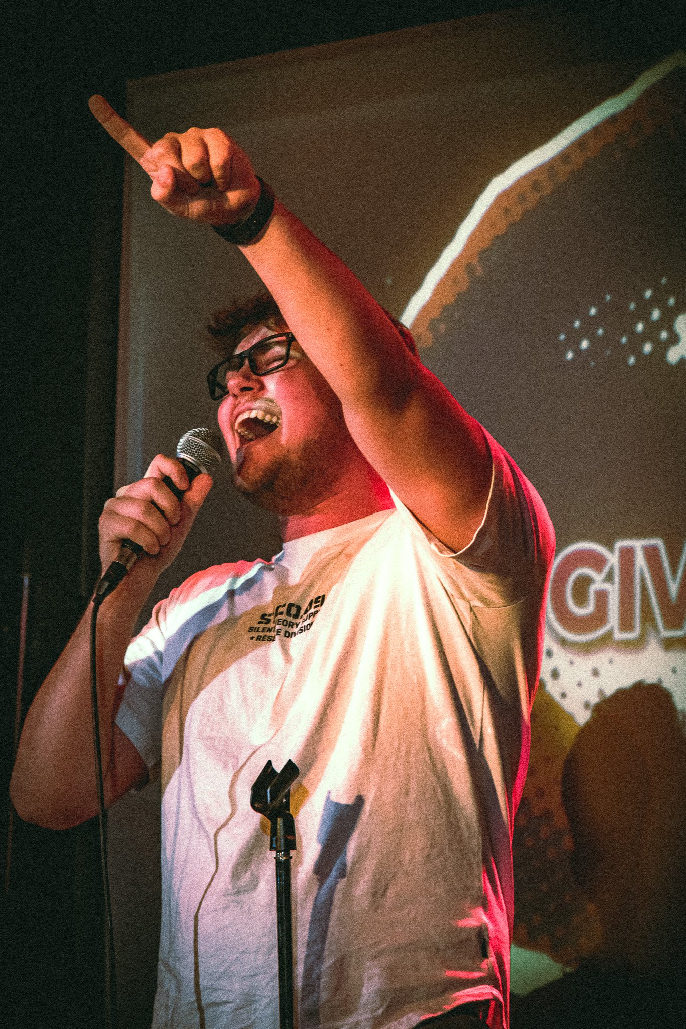 Man with glasses singing passionately into a microphone, raising his right arm with index finger pointed up. bandkaroke.live