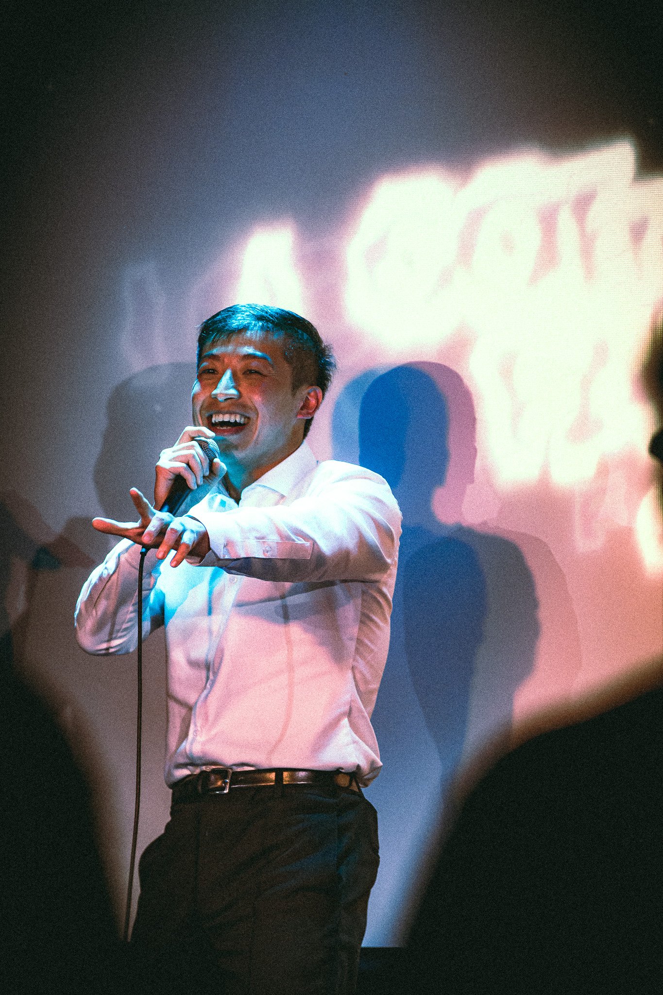Smiling man in white shirt performing stand-up comedy with a microphone on stage under colorful lights. bandkaroke.live
