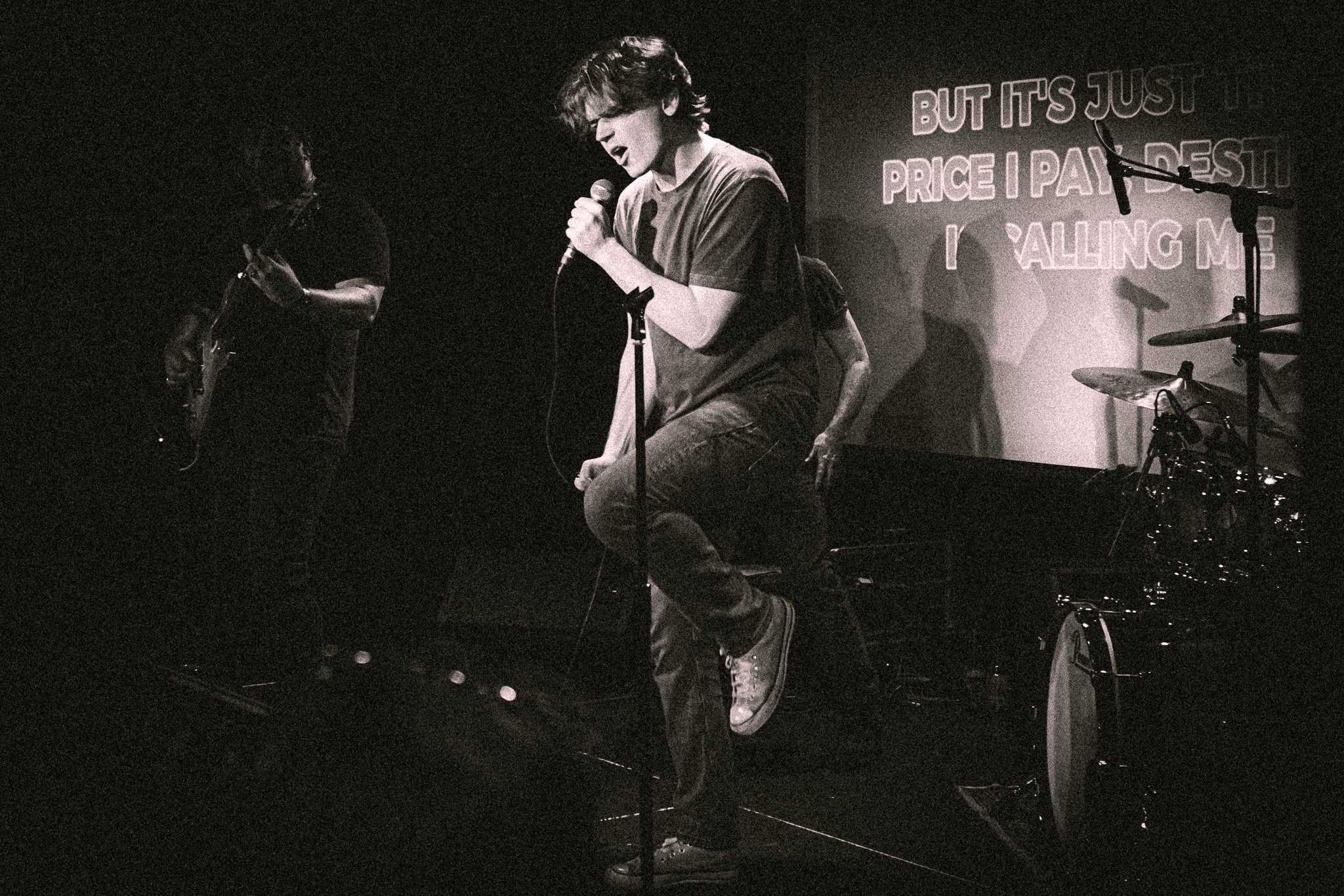 Black and white photo of a male singer holding a microphone on stage with a guitarist and drum set in the background. bandkaroke.live