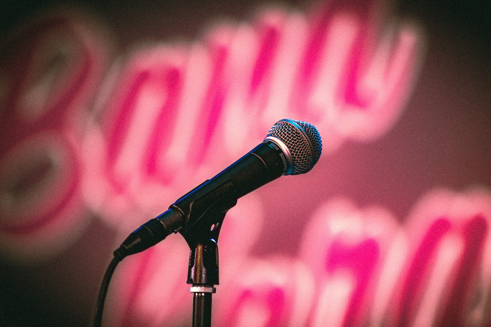 Close-up of a black Shure microphone on a stand with a blurred pink and red background. bandkaroke.live