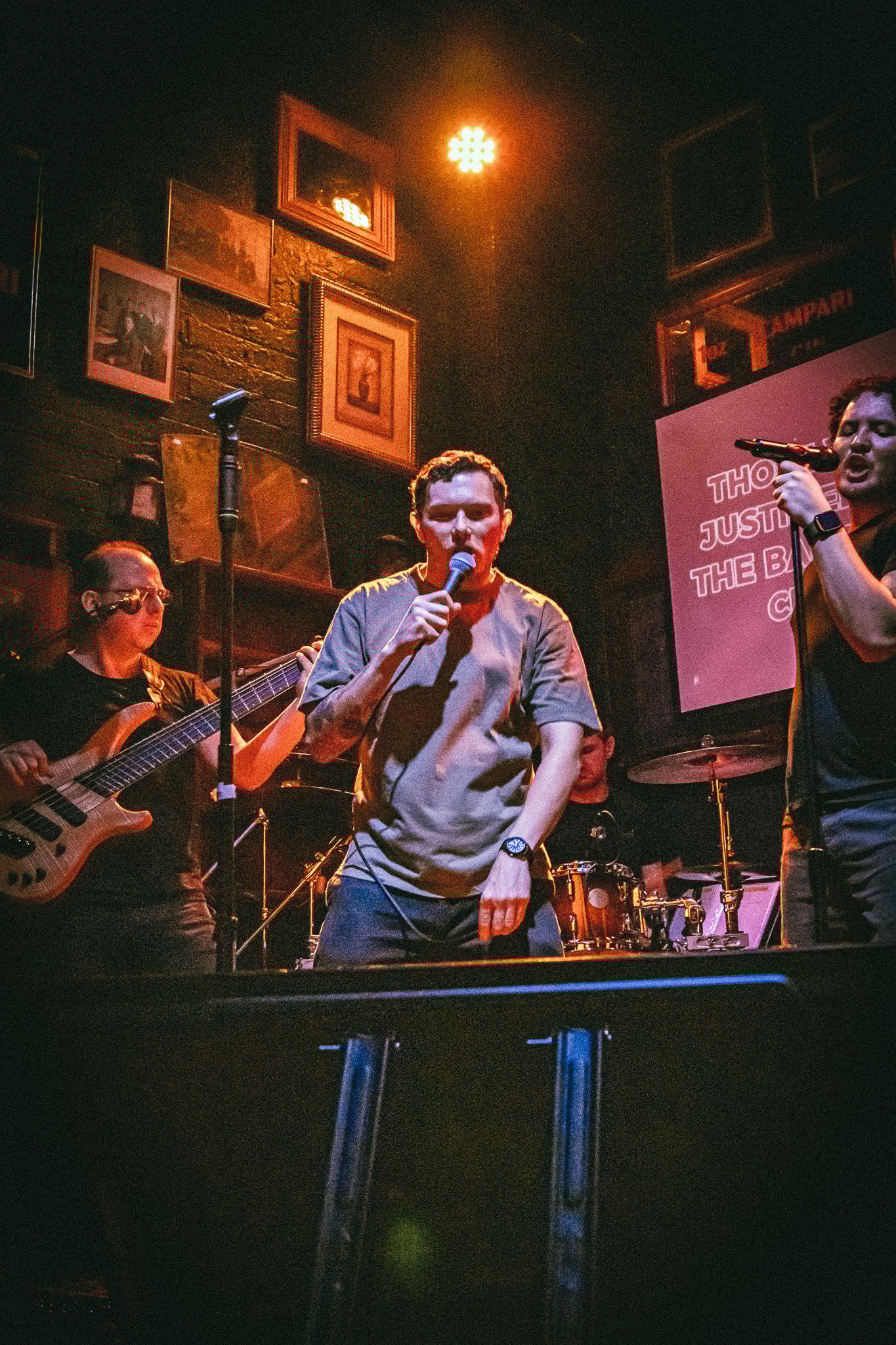 A band performing live in a dimly lit venue with a singer holding a microphone center stage, a bassist, and a drummer in the background. bandkaroke.live