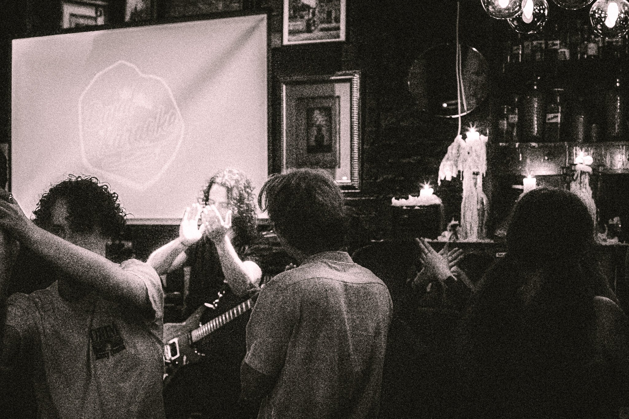 Grainy black-and-white photo of a group of people dancing and clapping in a dimly lit room with candles and framed pictures on the walls. bandkaroke.live