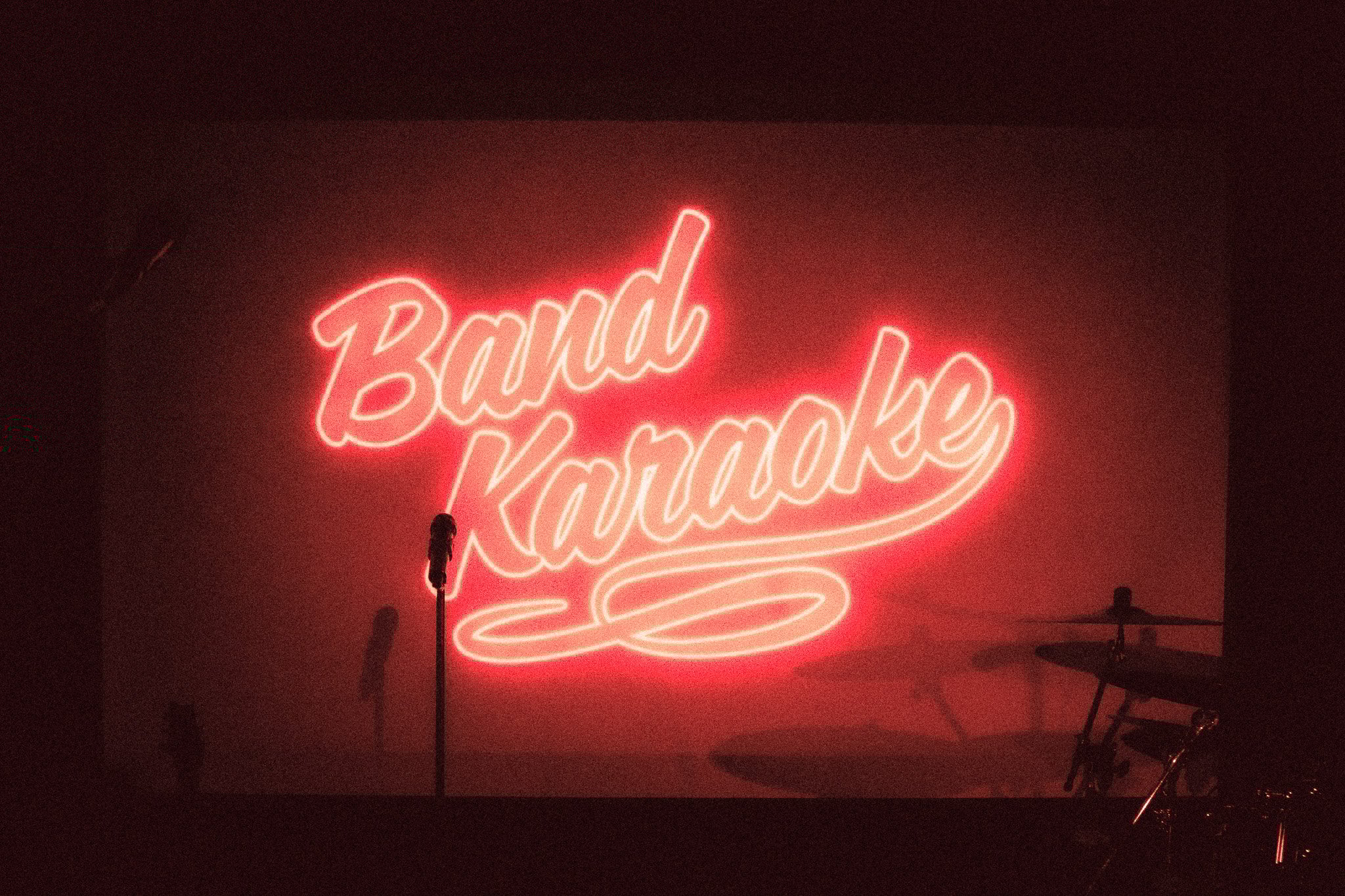 Microphone stand in front of a red neon sign reading 'Band Karaoke' with drum set partially visible in background. bandkaroke.live