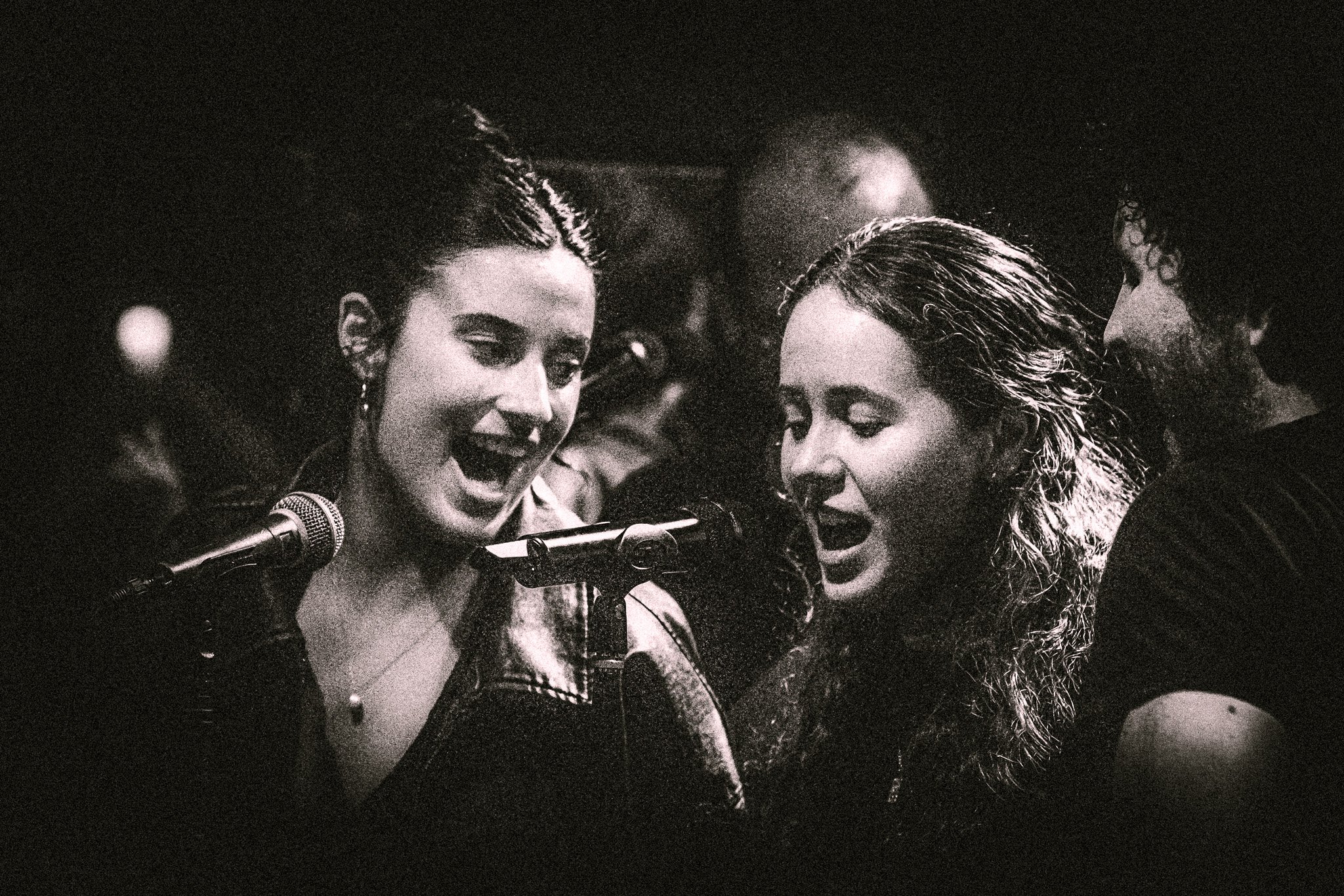 Black and white photo of two women singing into microphones with a man standing beside them in a dimly lit setting. bandkaroke.live