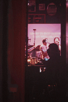 View through a doorway of a dimly lit band performing on stage, featuring a guitarist and singer with a visible microphone stand and drum set. bandkaroke.live