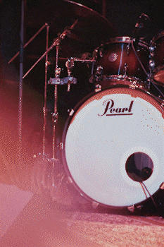 Close-up view of a Pearl bass drum with cymbals and drum stands in soft, colorful lighting.