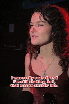 Curly-haired woman with subtle smile, lit by red light, with text overlay saying she was scared and shaking but had fun. bandkaroke.live