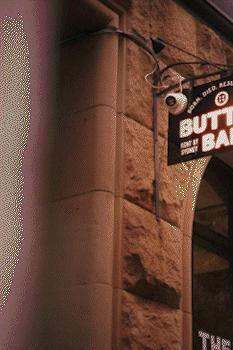 Security camera mounted on a textured brick wall above a sign reading 'BUTTON BAR'. bandkaroke.live