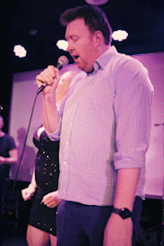 Man singing passionately into a microphone at a live event with a woman standing behind him. bandkaroke.live