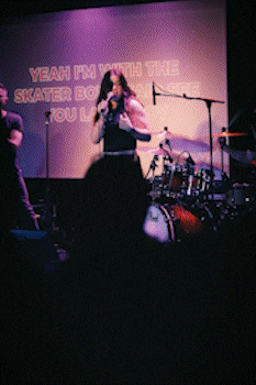 Female singer performing on stage with drummer and projected lyrics behind her during a live music event. bandkaroke.live