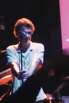 Person with glasses singing into a microphone on stage with dark background and purple lighting. bandkaroke.live