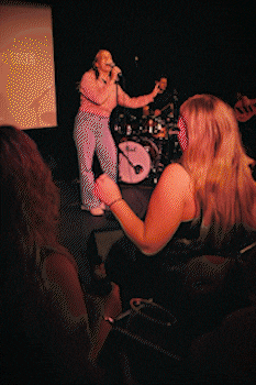 Female singer performing on stage with a drummer in the background and audience members watching closely. bandkaroke.live