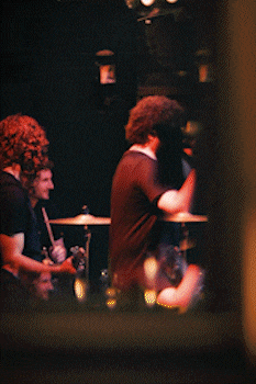 Three musicians performing on stage with a drummer, guitarist, and vocalist in dim orange lighting. bandkaroke.live