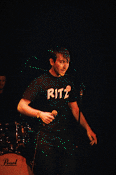 Male singer performing energetically on stage holding a microphone with a drummer and drum set in the background. bandkaroke.live