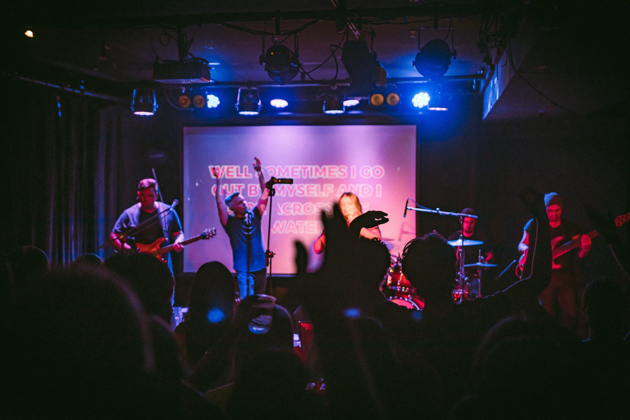 Band performing on stage with blue and purple lighting and audience silhouettes raising hands. bandkaroke.live