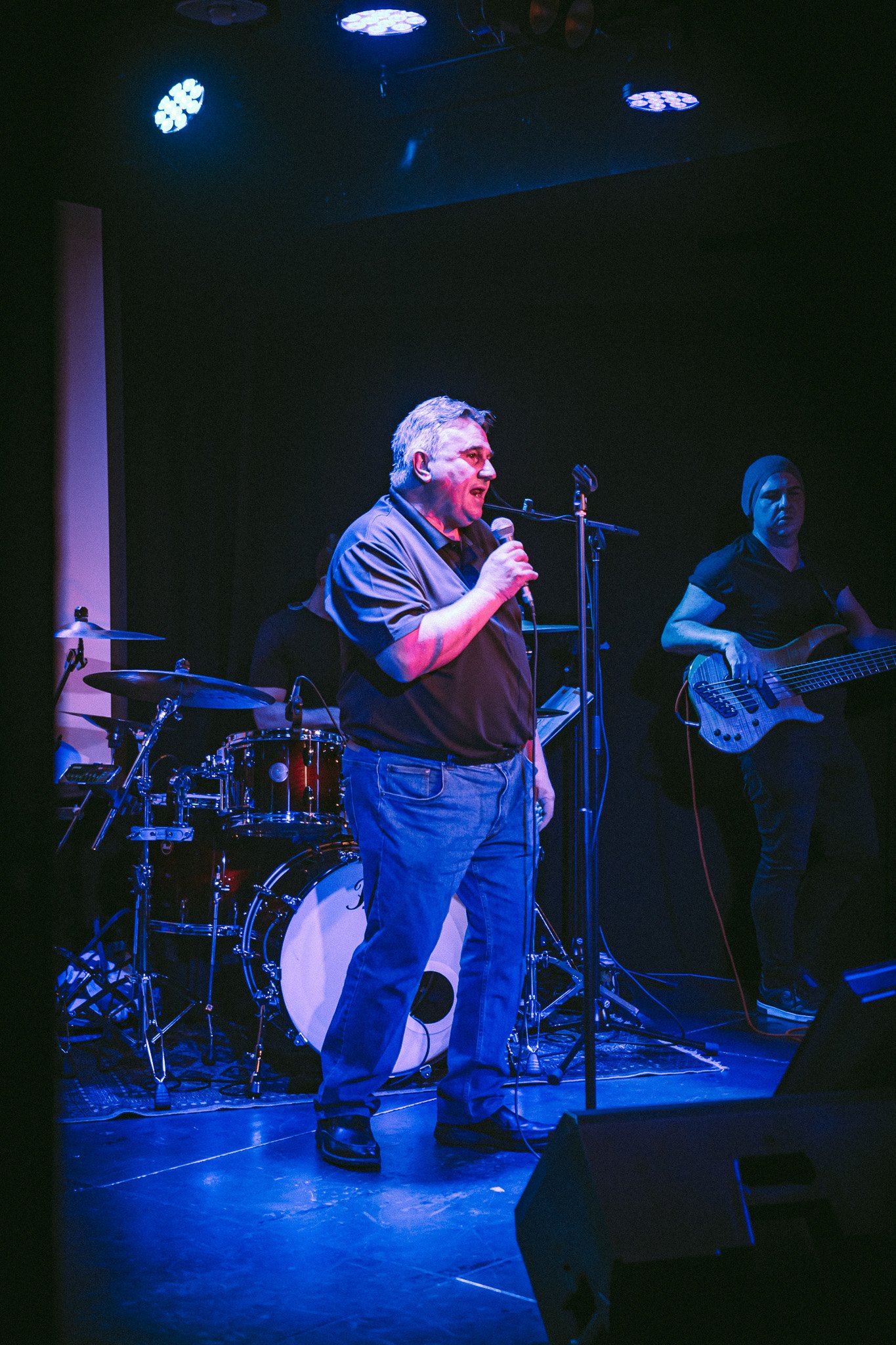 Man singing into a microphone on stage with a drummer and bass guitarist performing behind him under blue lighting. bandkaroke.live