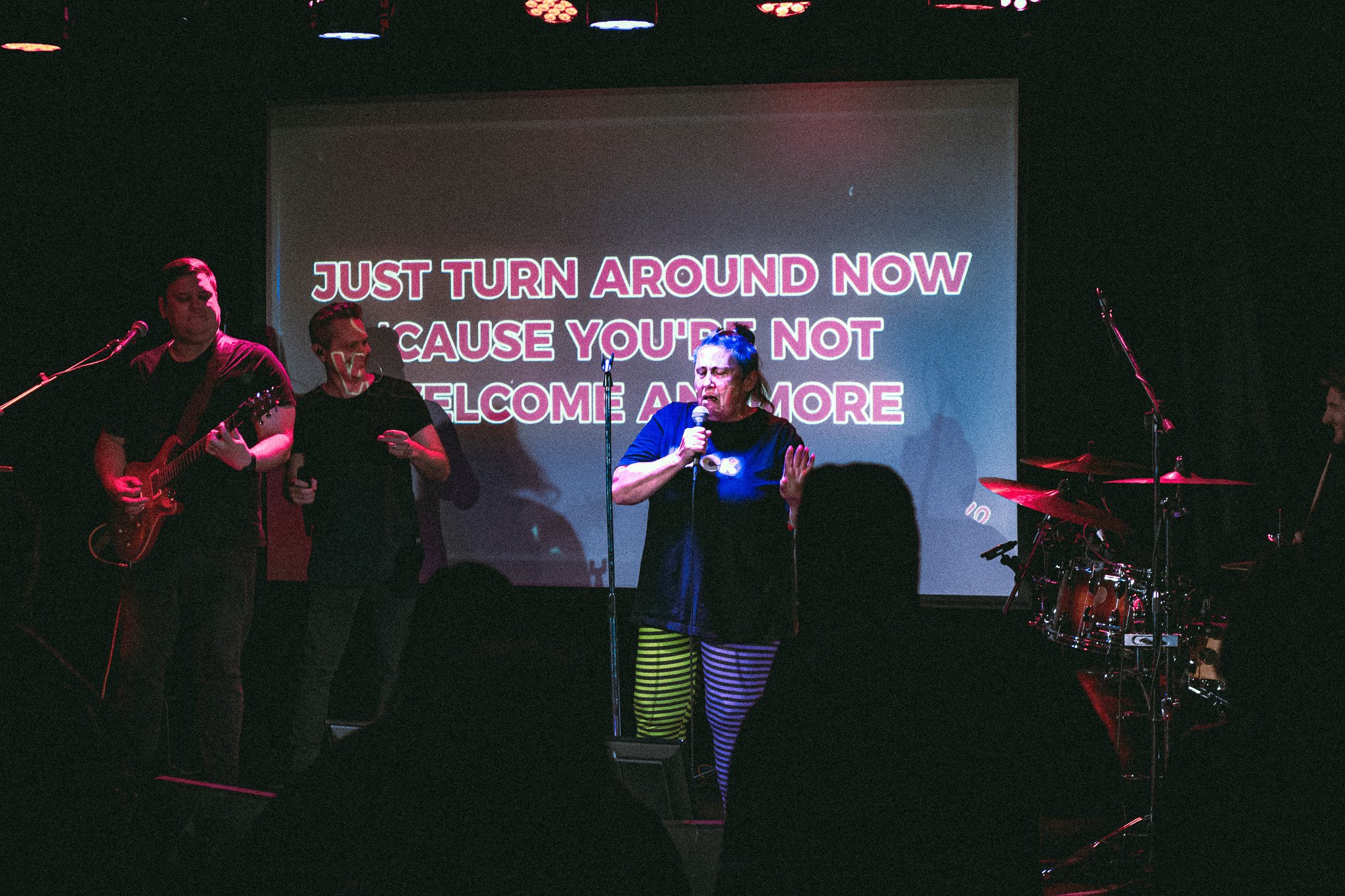 A woman singing on stage with a microphone, accompanied by a guitarist and a drummer, with karaoke lyrics projected behind her. bandkaroke.live