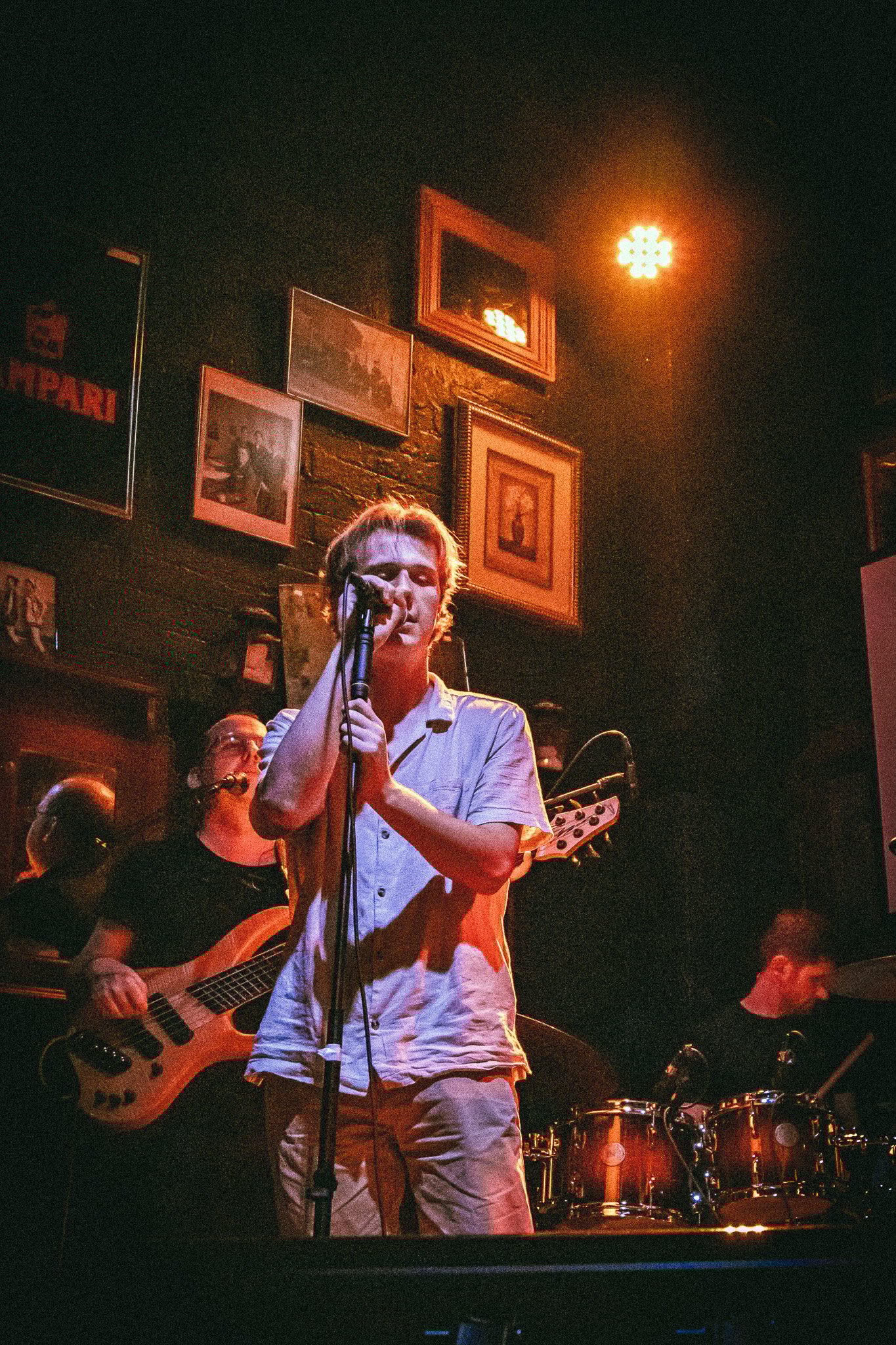 Male vocalist singing into a microphone with band members playing bass guitar and drums in a dimly lit venue with framed pictures on the wall. bandkaroke.live