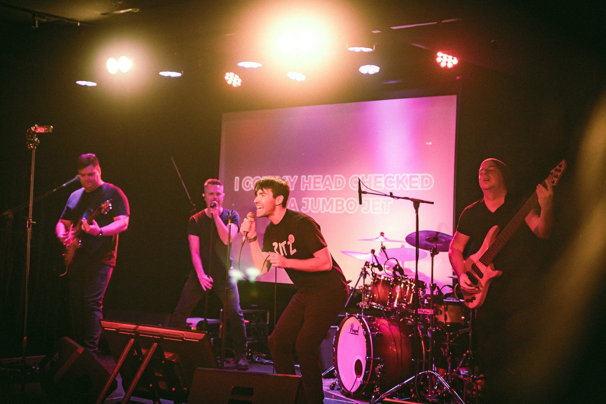 Band performing on stage with two vocalists, a guitarist, a bassist, and a drummer illuminated by stage lights. bandkaroke.live