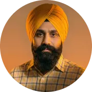 Man with a dark beard and mustache wearing a bright orange turban and a yellow checkered shirt against a warm beige background.