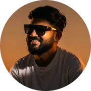Man with dark hair and beard wearing large black sunglasses and a light-colored shirt against a warm orange background.