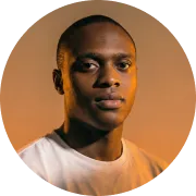 Portrait of a young Black man with a neutral expression, wearing a white shirt against an orange background.