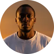Portrait of a serious young Black man with short hair wearing a white t-shirt against a warm-toned sky background.