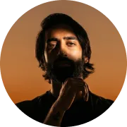 Portrait of a man with dark hair and beard, holding his chin thoughtfully against a warm, brownish-orange background.