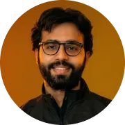 Portrait of a smiling man with a beard and mustache, wearing glasses and a black jacket, against an orange background.