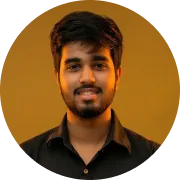 Portrait of a young man with dark hair and beard, wearing a black shirt, smiling against a warm orange background.