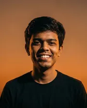 Portrait of a young man with short dark hair smiling against a warm gradient orange background.