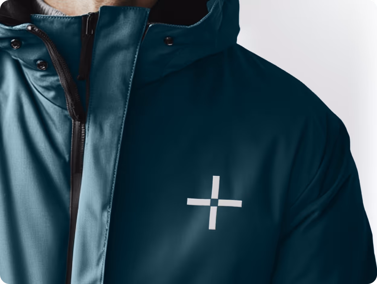 Close-up of a teal technical jacket featuring "The Be Company" plus-sign logo embroidered on the chest.