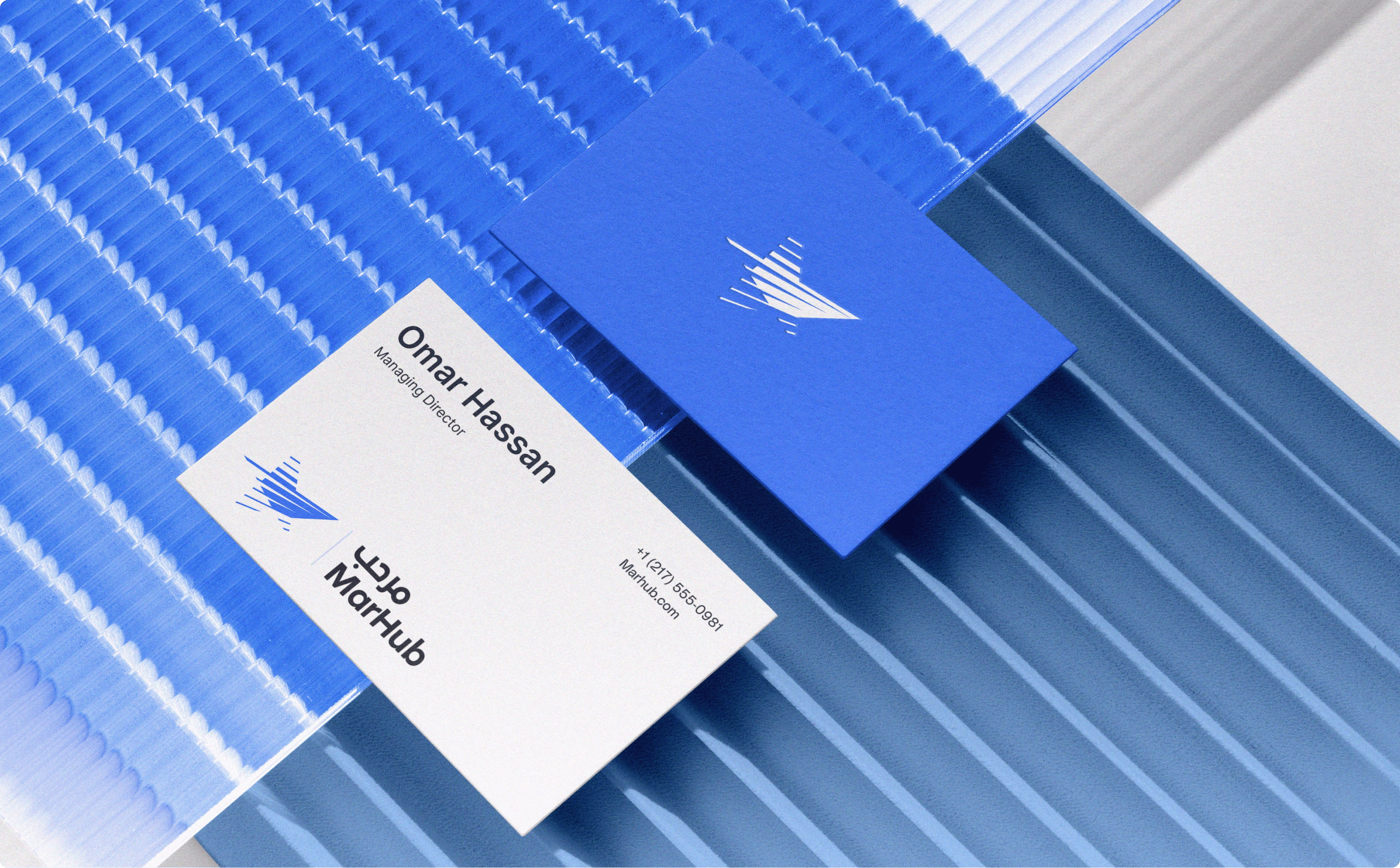 Marhub business cards for Omar Hassan on a blue ribbed surface, showing the ship logo and brand name. 