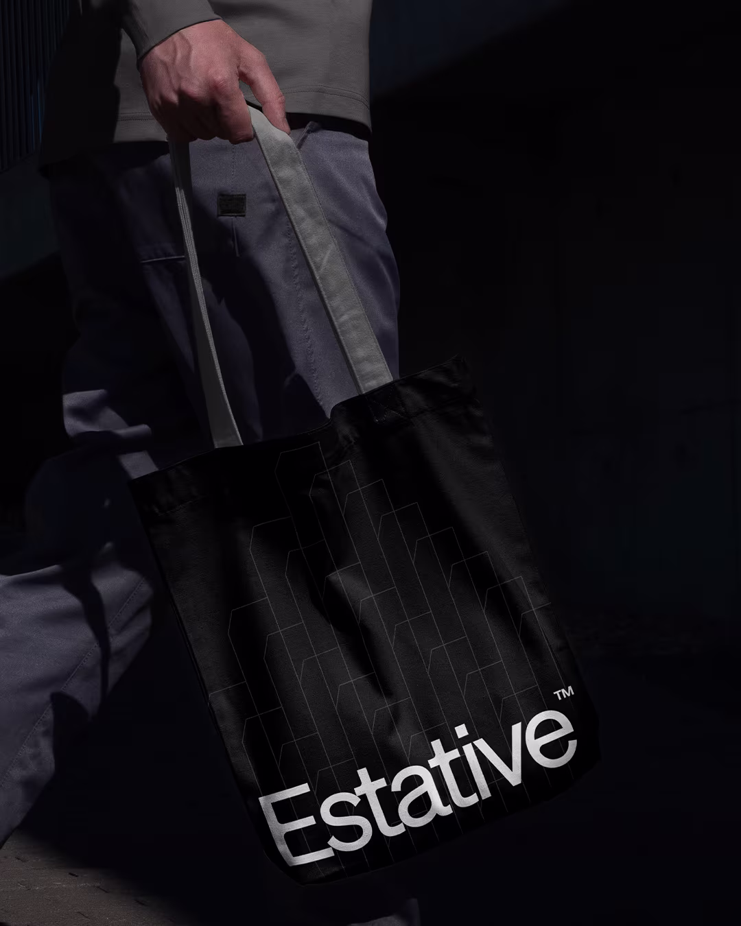 A person carrying a black Estative tote bag with a white wordmark and a subtle, geometric line pattern representing architectural silhouettes. 