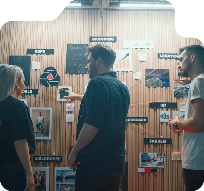 Three people standing and discussing in front of a wall covered with charts, images, and labels related to branding, marketing, and project planning.