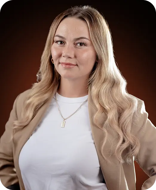 A smiling woman with long, wavy blonde hair, wearing a white shirt and a beige blazer, against a dark brown background.