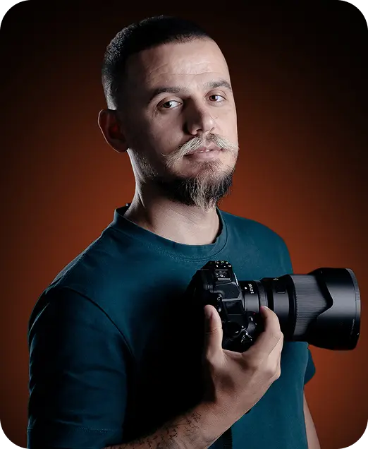 A man with a mustache and beard holding a camera against a dark orange background.