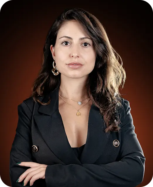 A confident woman with long, wavy hair wearing a black blazer, posing with her arms crossed against a dark brown background.
