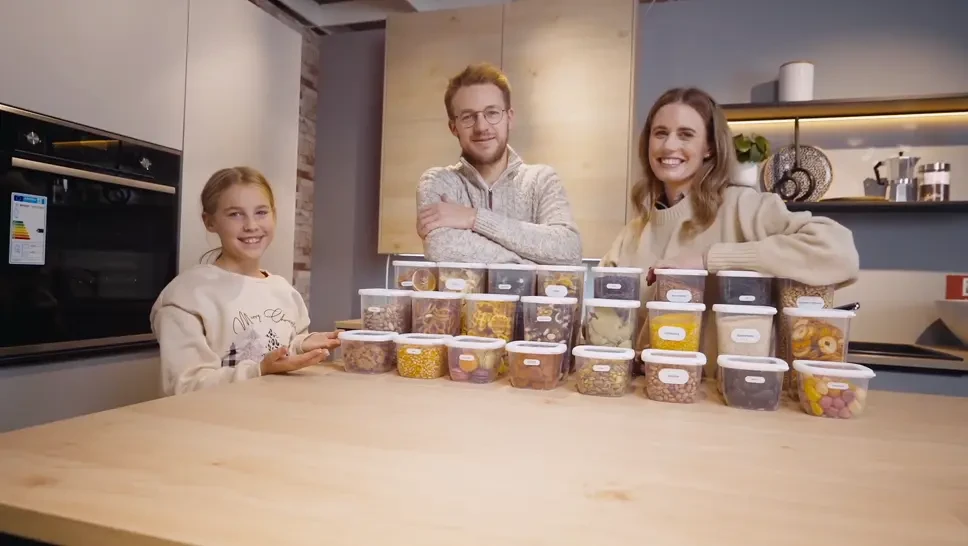 A smiling family of three, including a young girl, a man, and a woman, sitting behind a table filled with labeled clear plastic containers of assorted snacks in a modern kitchen.