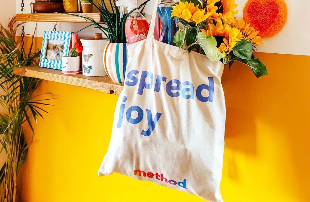 Method branded tote bag filled with sunflowers and household products hanging in a colorful home interior with bright yellow walls.