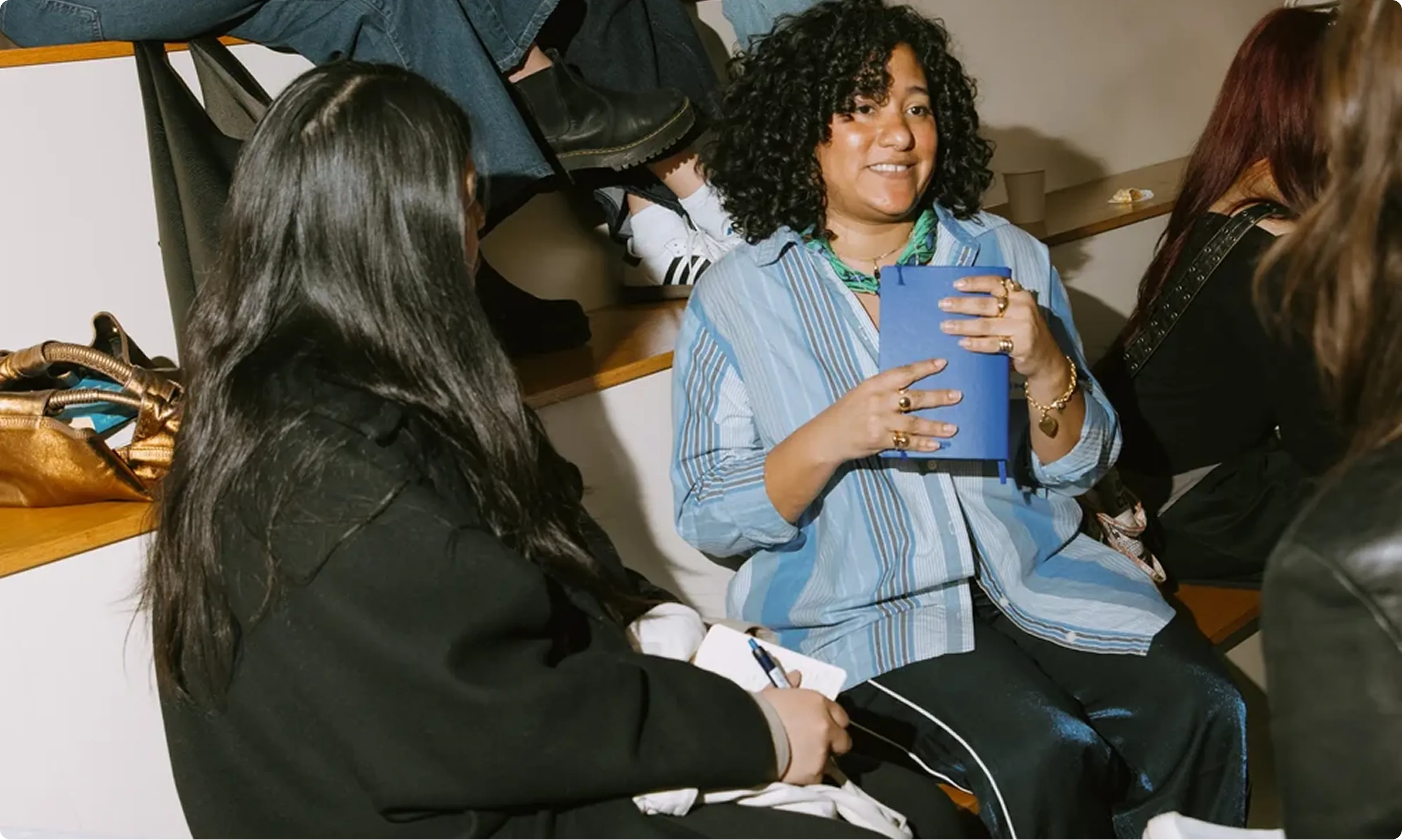 Two women sitting and talking indoors, one holding a blue notebook and the other writing in a notepad.
