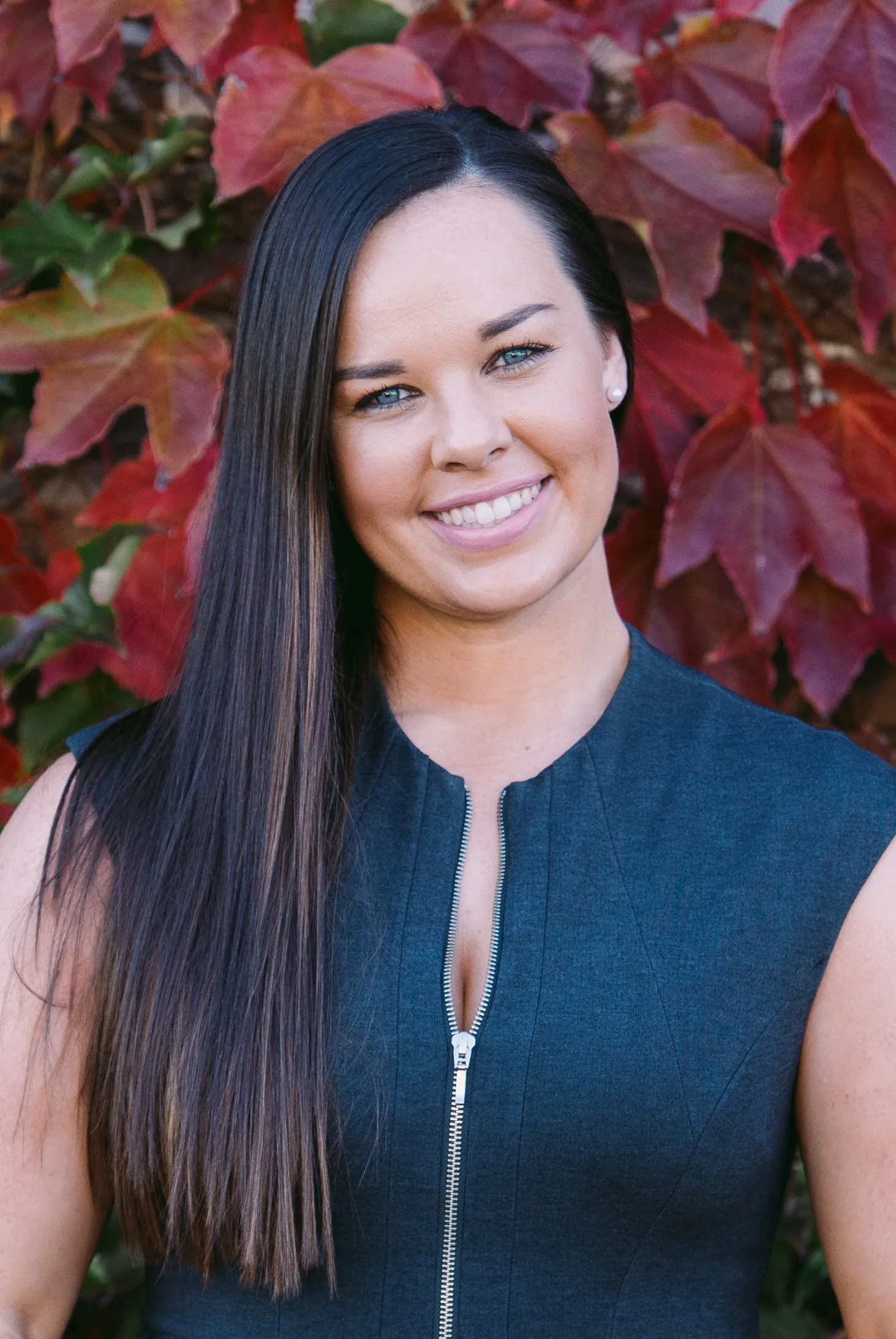 Smiling woman with long straight dark hair wearing a sleeveless zip-up top standing in front of red autumn leaves.