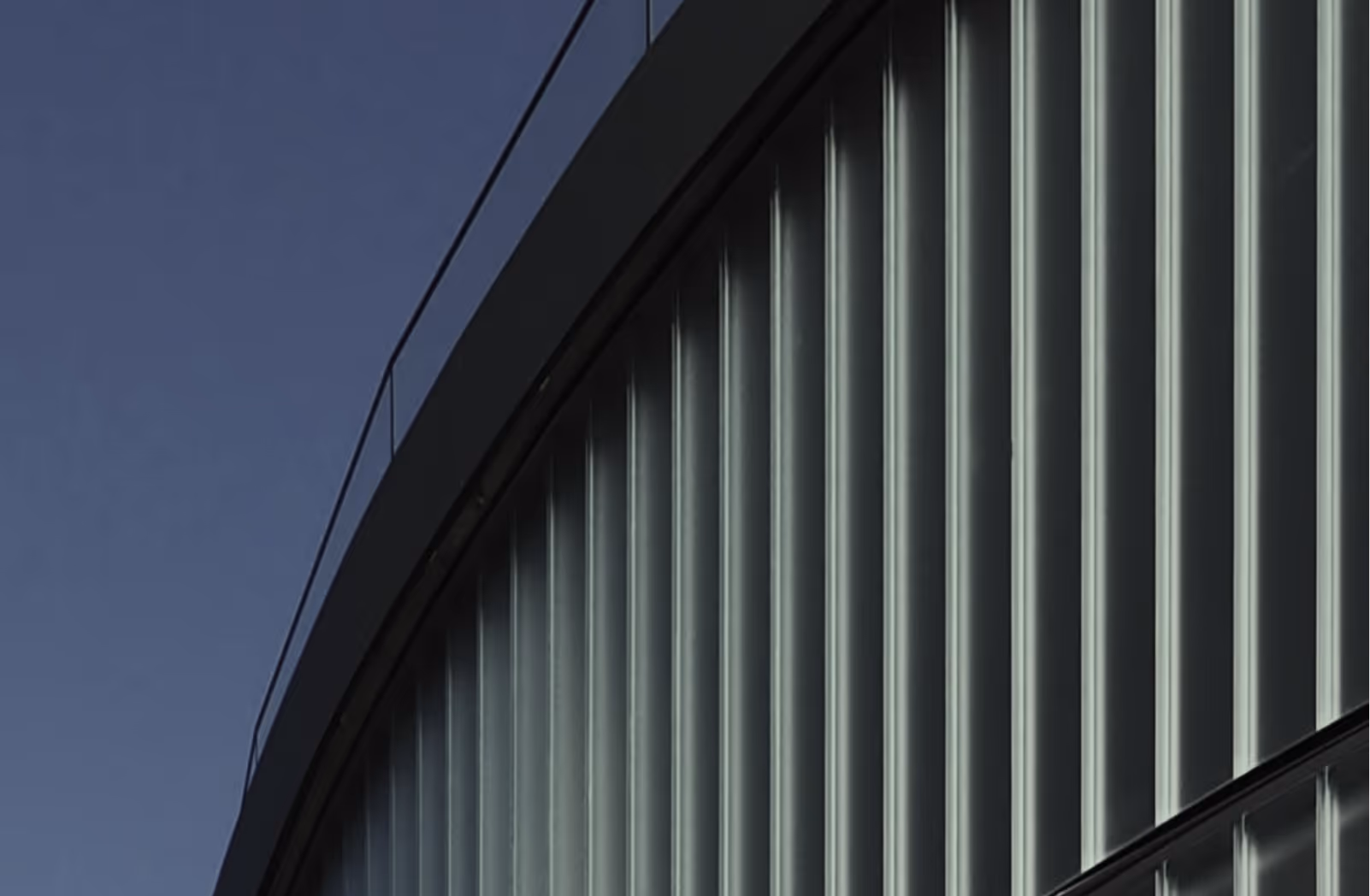 Fassade eines modernen Gebäudes mit vertikalen Glasfenstern und Dachgeländer vor blauem Himmel.