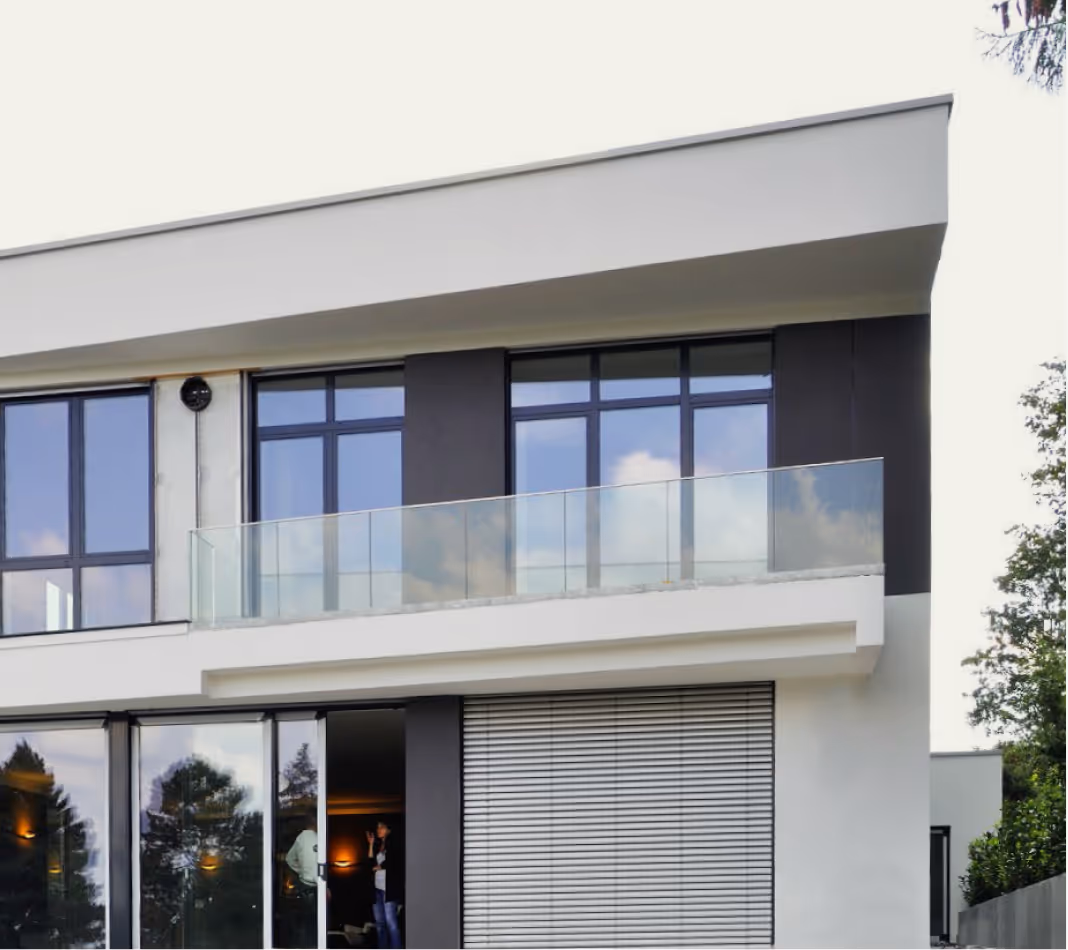 Modern zweistöckiges Haus mit großen Fenstern und Glasbalkon, umgeben von Bäumen.