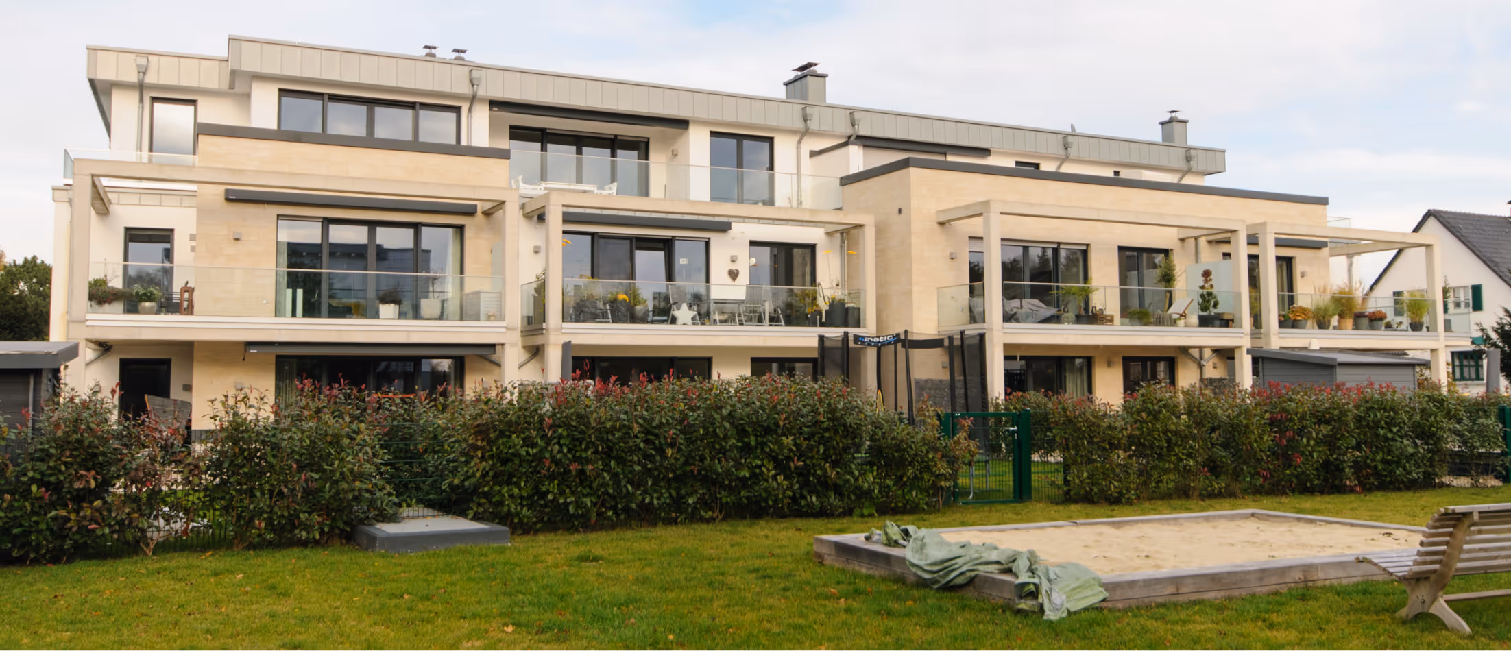 Modern residential building with balconies, hedge, green lawn, sandbox, and a wooden bench.