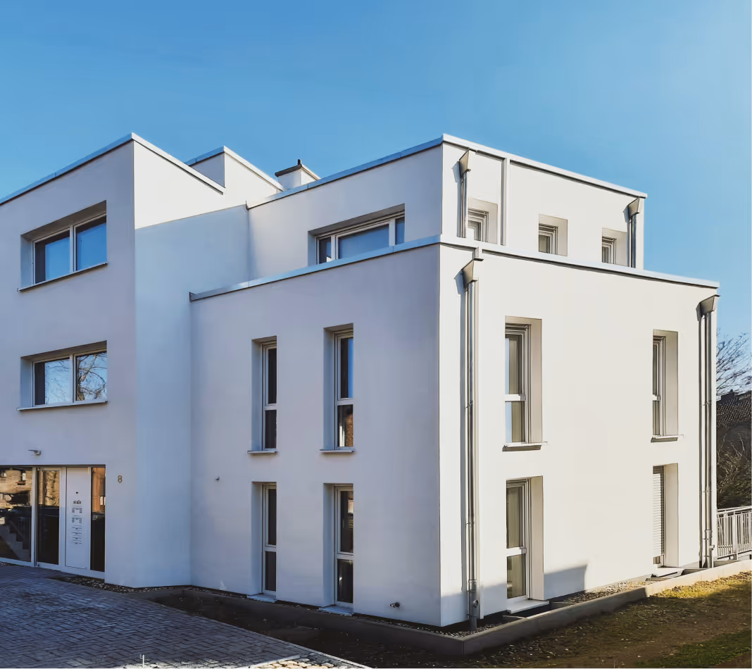 Modernes weißes Wohngebäude mit mehreren großen Fenstern unter klarem blauen Himmel.
