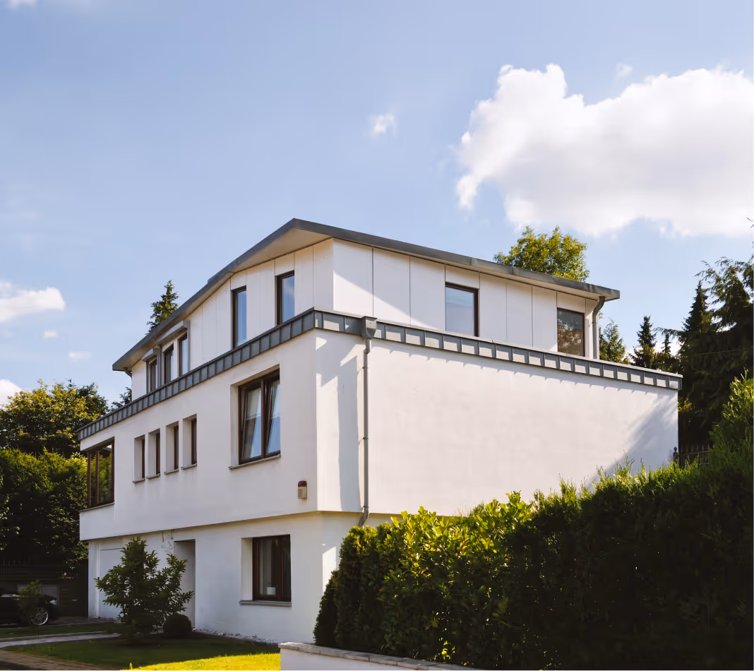Modern weißes Wohnhaus mit mehreren Fenstern und grünem Vorgarten unter blauem Himmel.